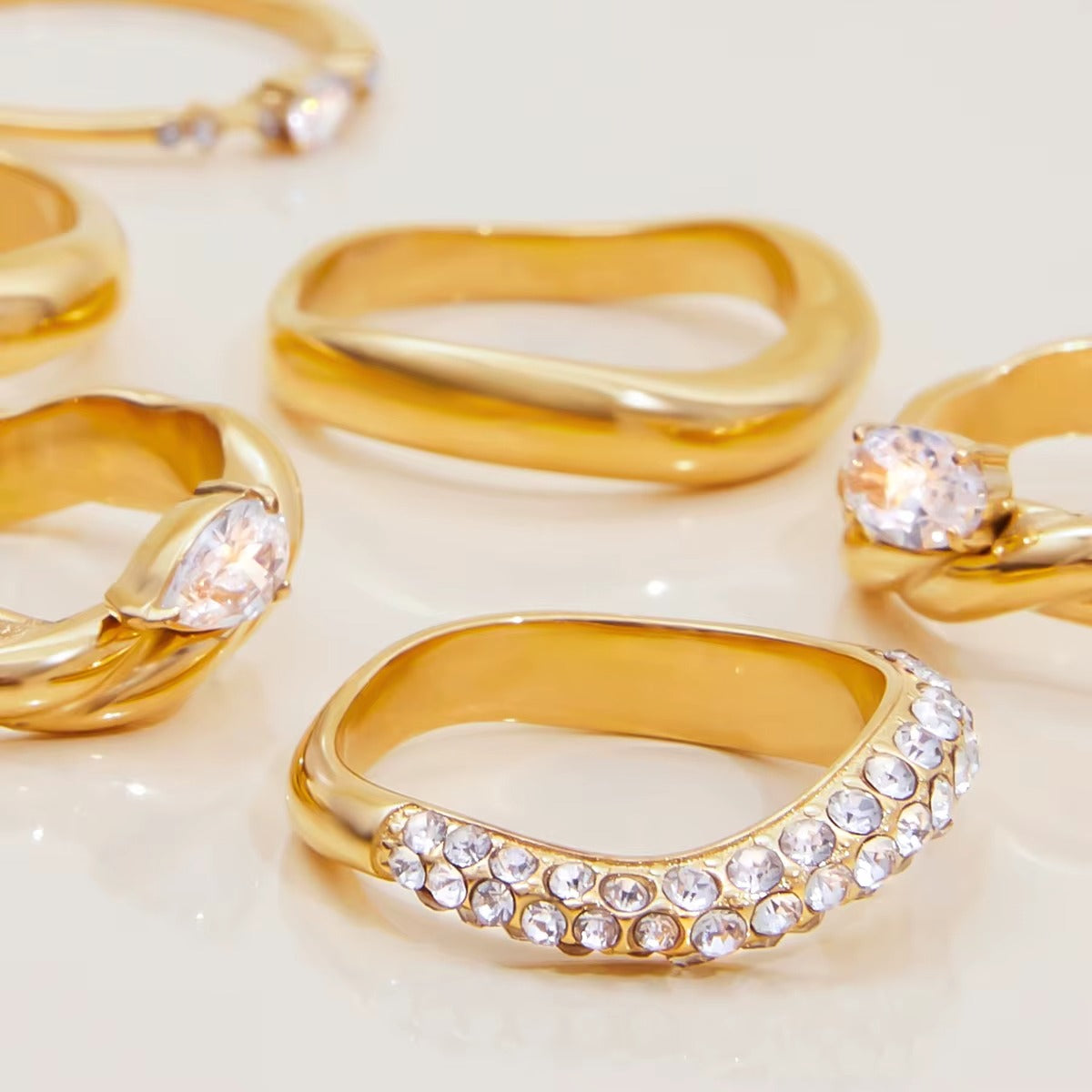 Several gold Misbehave rings scattered on a light surface, showing wavy, pavé and stone-set band styles from the collection up close.
