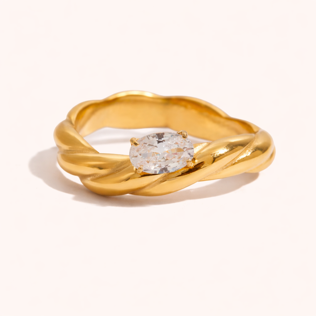 Twisted gold rope-band Misbehave RING set with a single oval clear stone, shown on a cream backdrop in studio.
