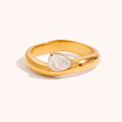 Smooth wavy gold Misbehave RING set with a single pear-shaped clear stone, shown on a cream backdrop in studio.