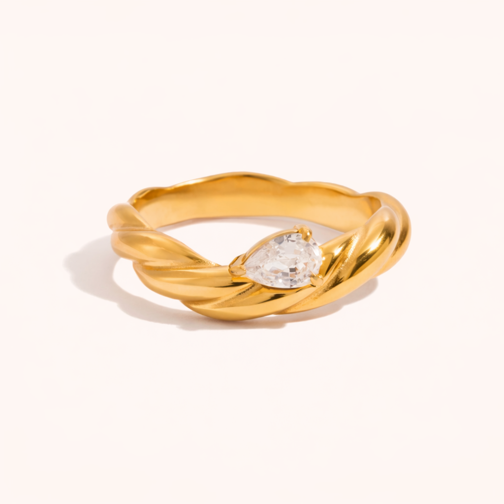 Twisted gold rope-band Misbehave RING set with a single pear-shaped clear stone, shown at an angle on a cream backdrop.