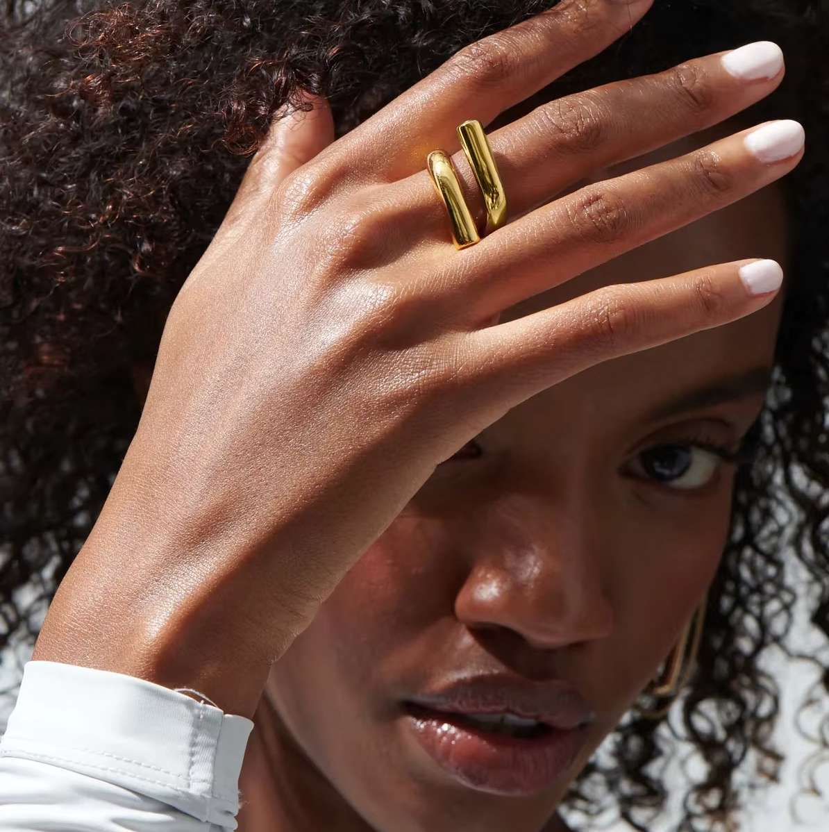 Model wears the gold Mischa RING with its parallel-bar design, hand raised to shield her face.