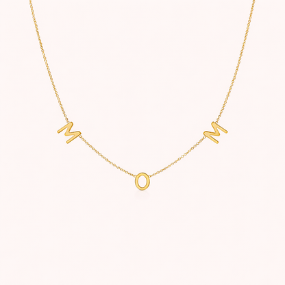 The Mom NECKLACE in gold, individual letters spelling MOM dangling from a fine chain, shown on a cream background.