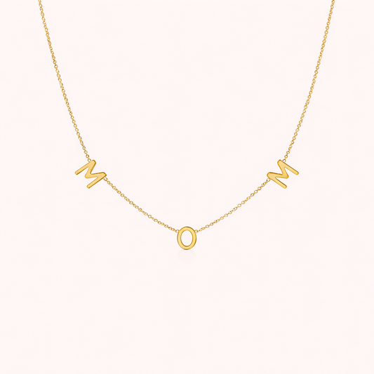 The Mom NECKLACE in gold, individual letters spelling MOM dangling from a fine chain, shown on a cream background.