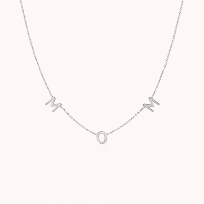 The Mom NECKLACE in silver, individual letters spelling MOM dangling from a fine chain, shown on a cream background.