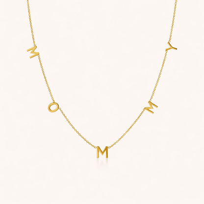 The Mommy NECKLACE in gold, individual letters spelling MOMMY dangling from a fine chain, shown on a cream background.