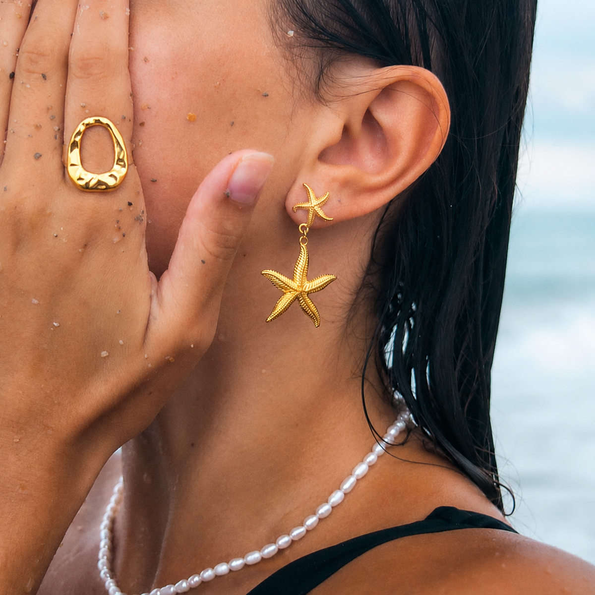 Model wears the gold Move Over RING on a sandy hand raised to her cheek, a starfish earring and pearl necklace visible nearby.