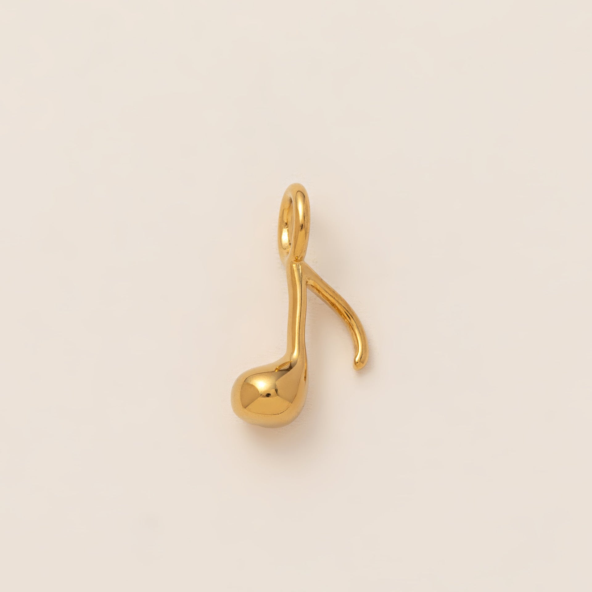 The Music Note CHARM in gold, an eighth note pendant with a polished finish, shown in close-up studio photography on cream.