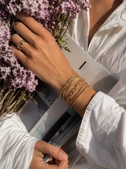 Gold chain bracelets worn stacked on the wrist with a ring, styled near a bouquet of lilac flowers, Folie a Trois bracelet collection.