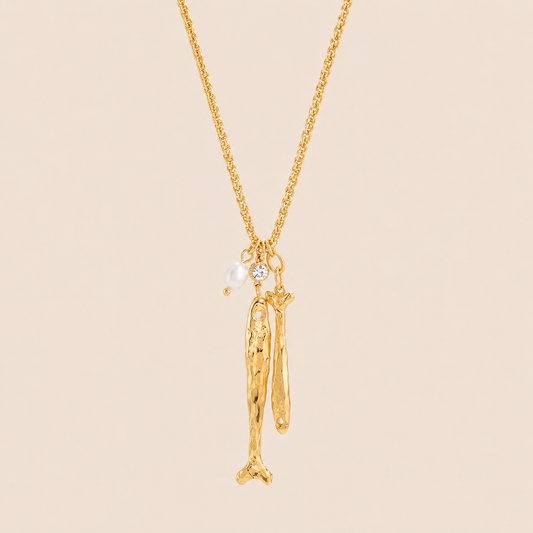 Gold Naiade NECKLACE with two textured fish-bone charms and a pearl and crystal accent, laid flat on a beige background.