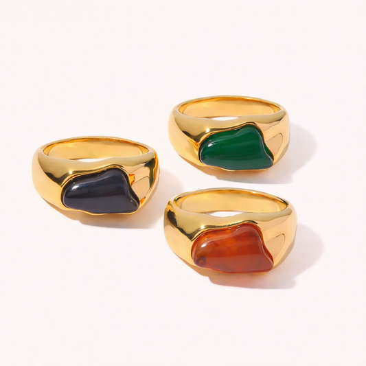 Three gold signet-style New Fashion RING styles with irregular cabochon stones in black, green and amber, on a cream backdrop.