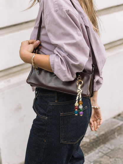 Folie à Trois Nodo BAG CHARM, a multicolor beaded strand on cream cord, clipped to a brown leather shoulder bag worn over denim.
