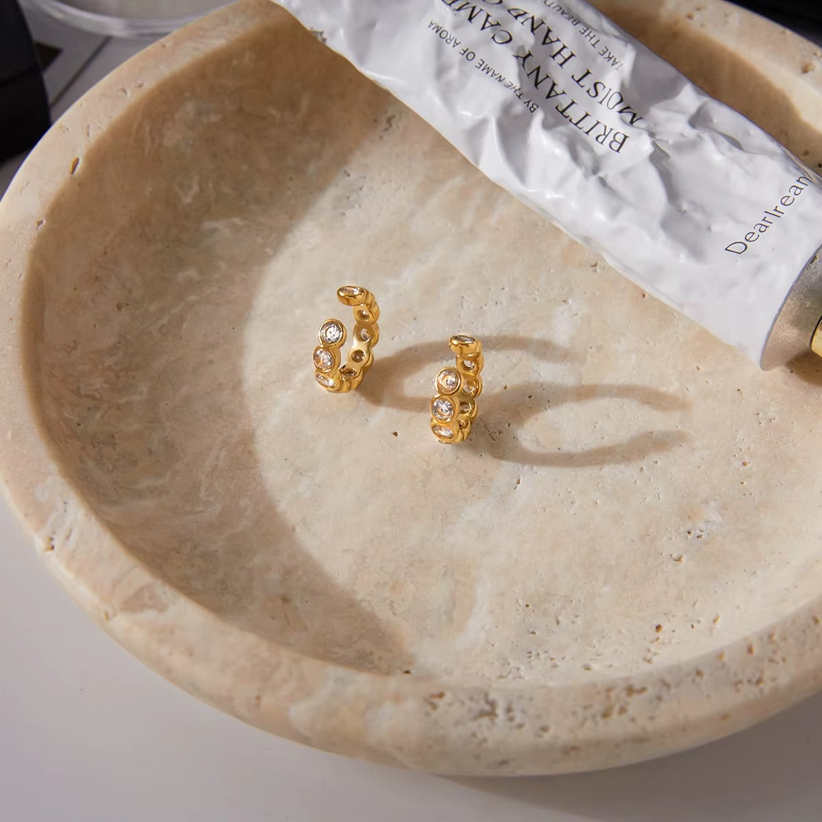 Pair of Nomad ear cuffs laid in a stone bowl beside a hand cream tube, showing the crystal-lined gold bands up close.