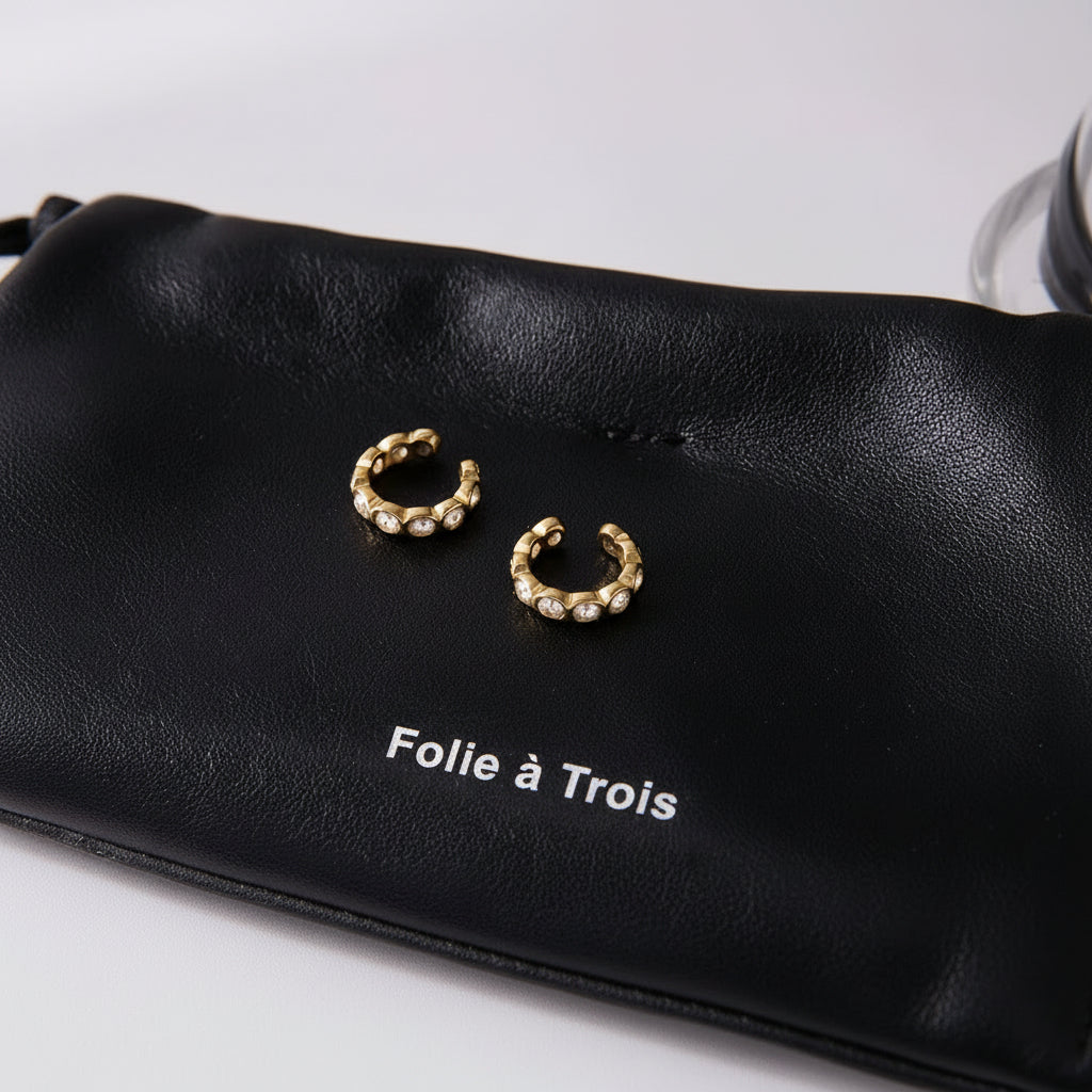 Pair of Nomad ear cuffs resting on a black pouch printed with the Folie a Trois logo, crystals catching the light.