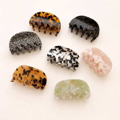 Seven different acetate hair claw clips in tortoiseshell, leopard, mottled and marble patterns laid out together on a cream background.