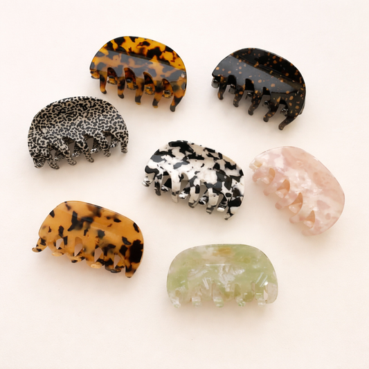 Seven different acetate hair claw clips in tortoiseshell, leopard, mottled and marble patterns laid out together on a cream background.