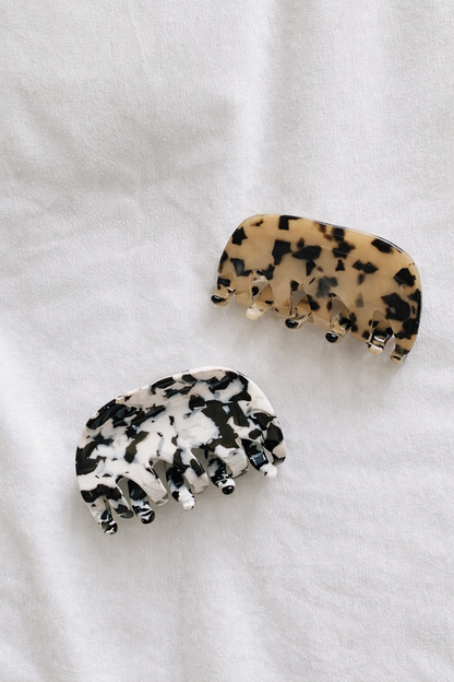 Two acetate hair claw clips, one tan tortoiseshell and one black and white mottled, resting together on white fabric.