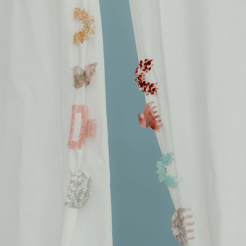 An assortment of seven small acetate hair clips in flower and butterfly shapes hanging on sheer curtains.