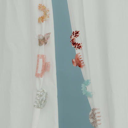 An assortment of seven small acetate hair clips in flower and butterfly shapes hanging on sheer curtains.