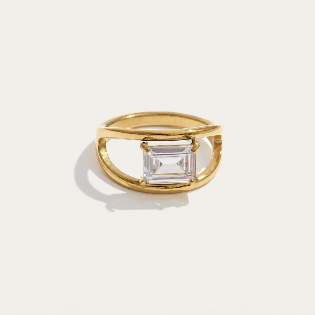Gold open-band ring with an emerald-cut white zirconia set between split shoulders on a cream backdrop — Folie à Trois Ovale RING