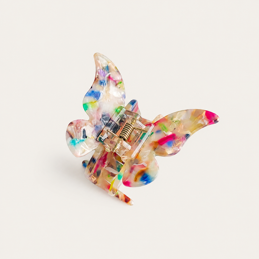 Papillon HAIR CLIP butterfly-shaped claw clip in translucent resin with multicolored paint splatters, on beige.