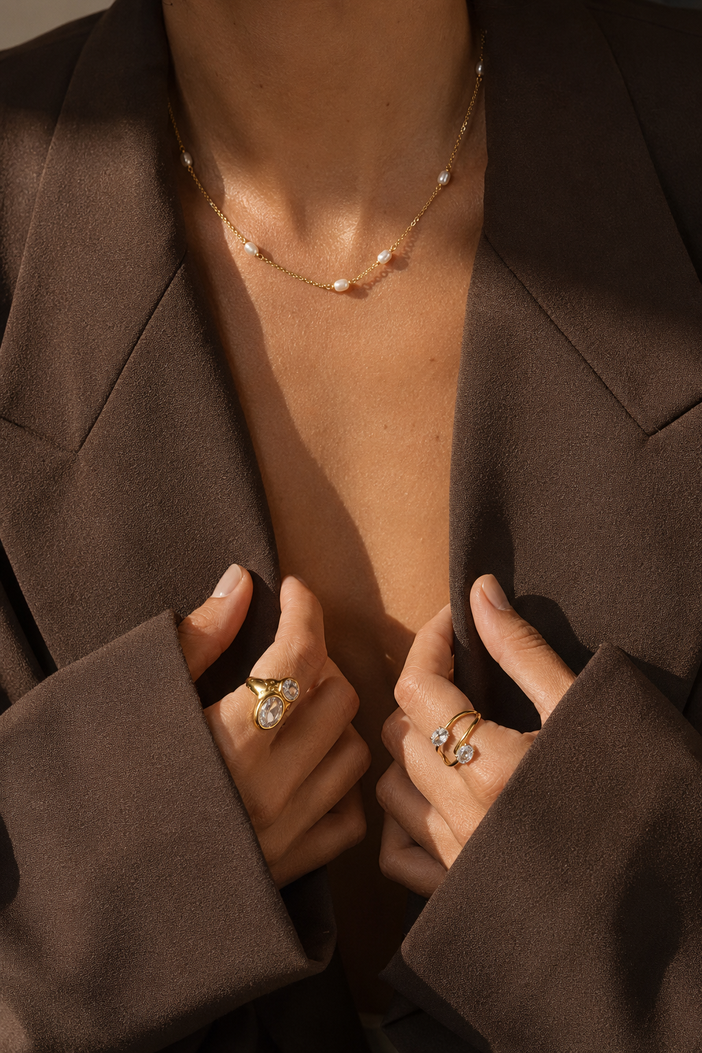 Model wears the gold Perle NECKLACE with its pearl stations at the neckline, hands resting on a brown blazer.