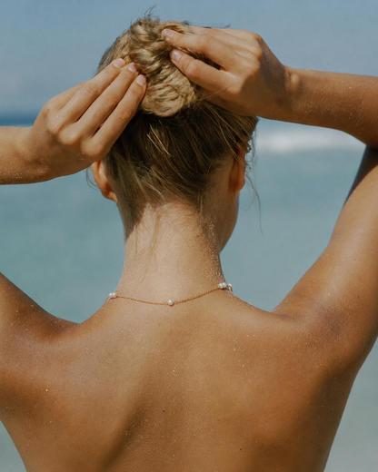 Model wears the gold Perle NECKLACE with its pearl stations visible on the back of the neck, hair pulled up at the beach.