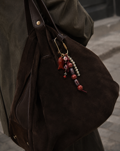 Folie à Trois Pescado BAG CHARM in burgundy and purple beaded strands with a leaf-shaped enamel charm, clipped to a dark suede bag.