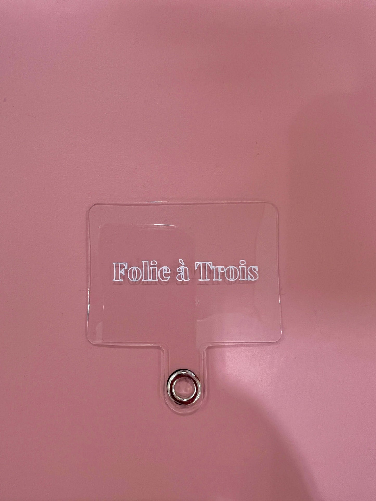 A clear acrylic phone cord adapter tag embossed with the Folie à Trois logo, with a small metal grommet ring, on a pink surface.
