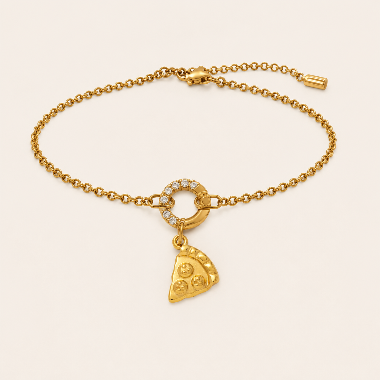A gold chain bracelet with a small ring set with clear stones and a dangling pizza slice charm, displayed on a light cream background.