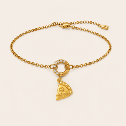 A gold chain bracelet with a small ring set with clear stones and a dangling pizza slice charm, displayed on a light cream background.