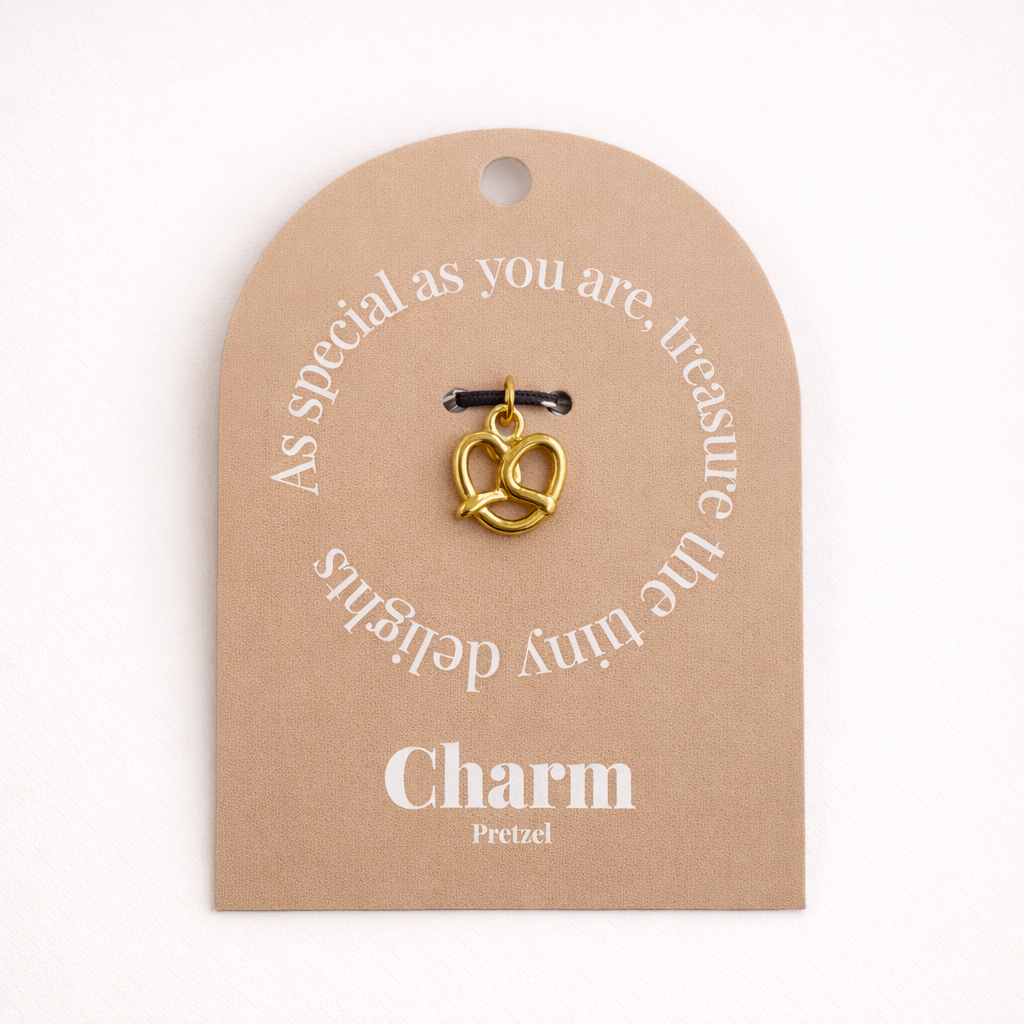 The Pretzel CHARM displayed on a tan Charm gift tag, the gold pretzel pendant hanging from a black cord.