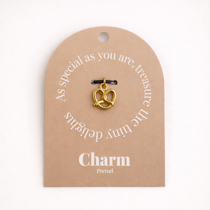 The Pretzel CHARM displayed on a tan Charm gift tag, the gold pretzel pendant hanging from a black cord.