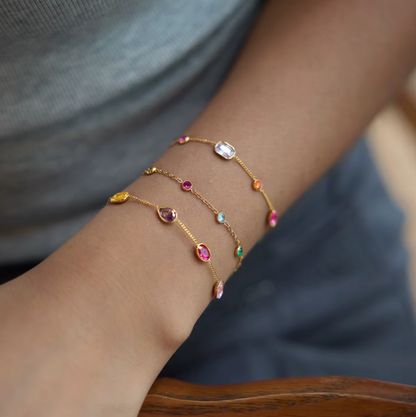 Arm resting on a wooden chair wearing three stacked gold Riva BRACELETs with multicolored oval, round and emerald-cut stones.