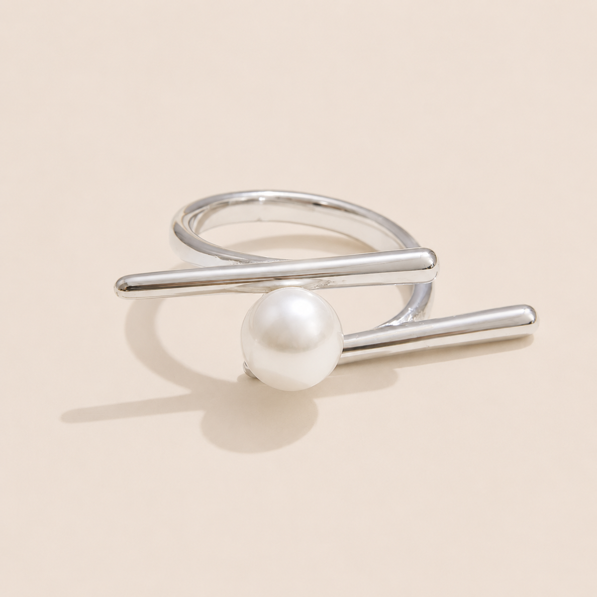 Silver double-band ring with two crossing bars cradling a single round pearl, shown on a cream backdrop — Folie à Trois Riviera Club RING.