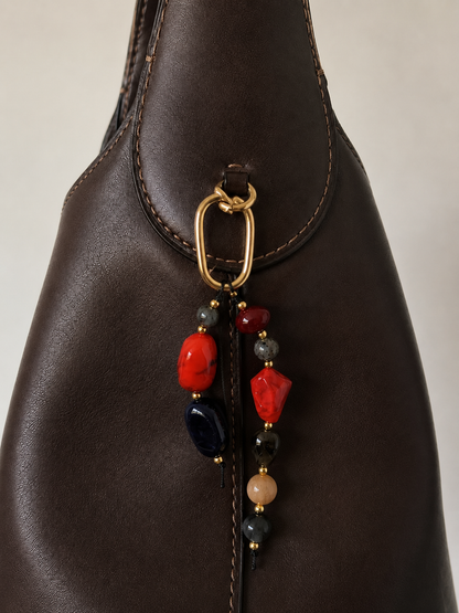 Folie à Trois Rocca BAG CHARM in red, black and grey natural stone beads, clipped to a dark brown leather bucket bag.