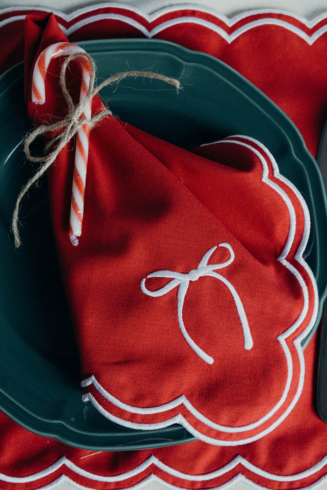 Red scalloped napkin wrapped around a candy cane with twine, set on the matching Rosie Scallop placemat with bow and a dark plate.