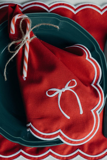 Red scalloped napkin wrapped around a candy cane with twine, set on the matching Rosie Scallop placemat with bow and a dark plate.