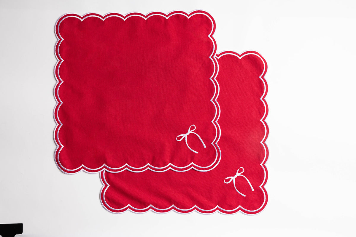 Flat lay of two folded red napkins with white scalloped trim and an embroidered bow, the Rosie Scallop napkins with bow set of two.