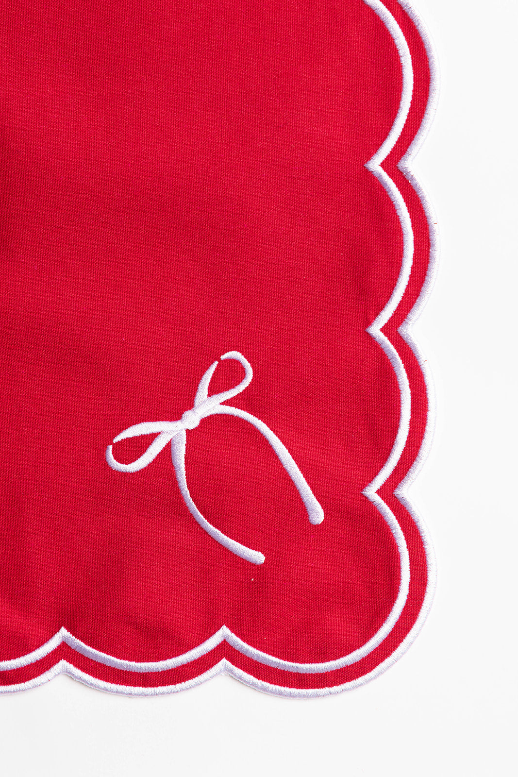 Close-up of a red napkin corner with white scalloped trim and a delicate embroidered bow, from the Rosie Scallop napkins with bow.