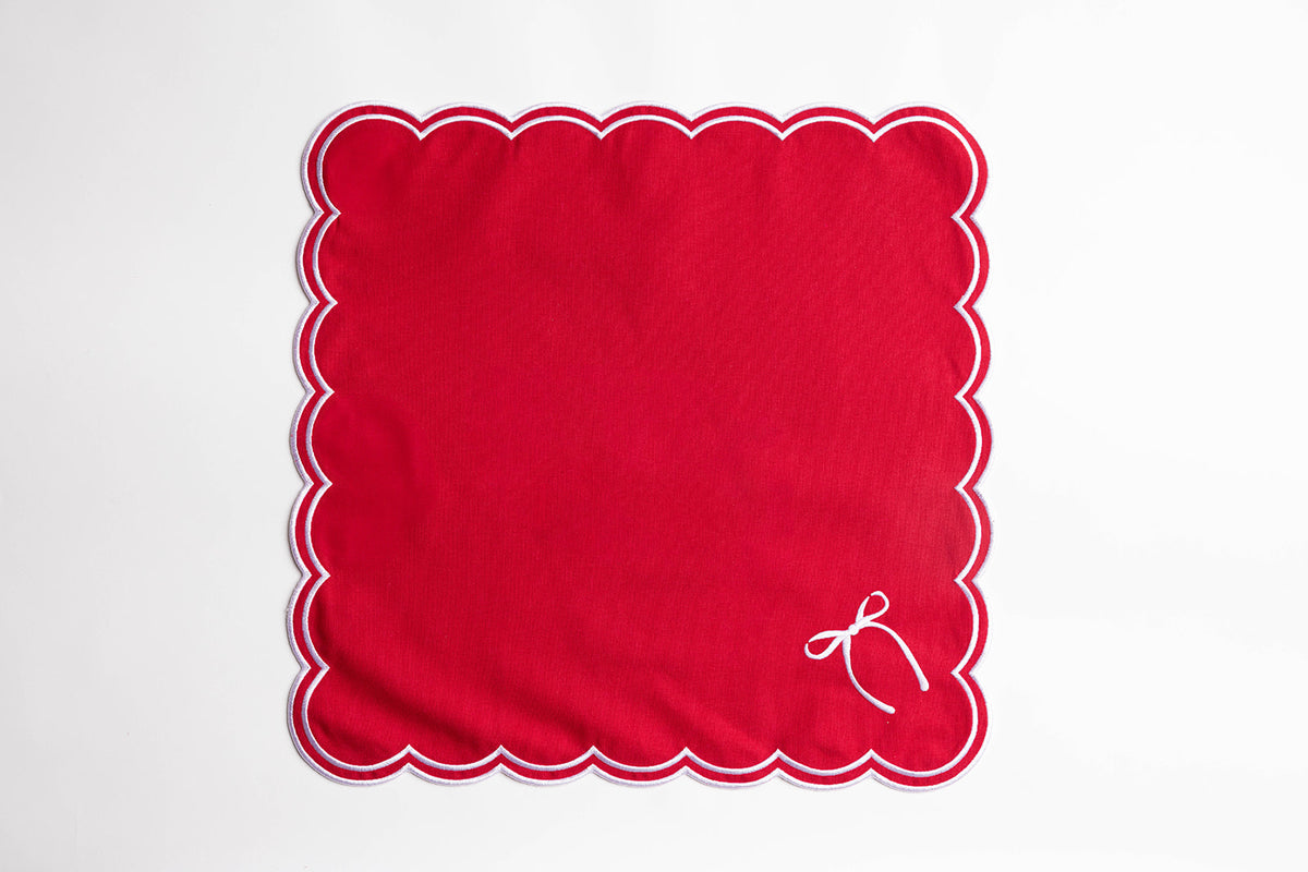 Flat lay of a single red square napkin with white scalloped edging and an embroidered bow, the Rosie Scallop napkin design.