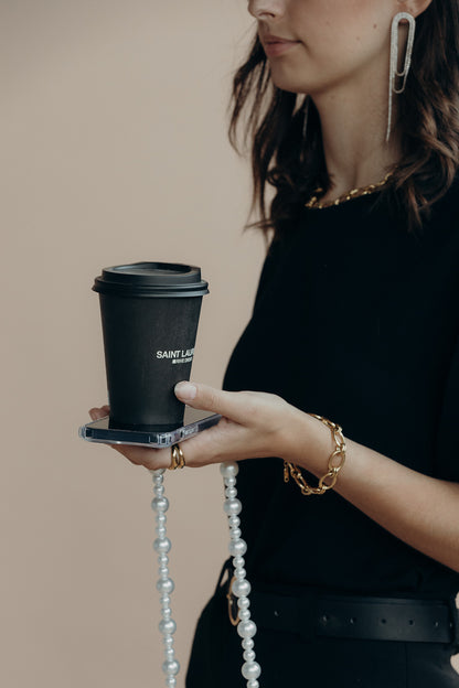 Model holding a phone with the Folie à Trois Sabine Pearl wholesale phone chain draped from it, paired with a gold chain bracelet and a coffee cup.