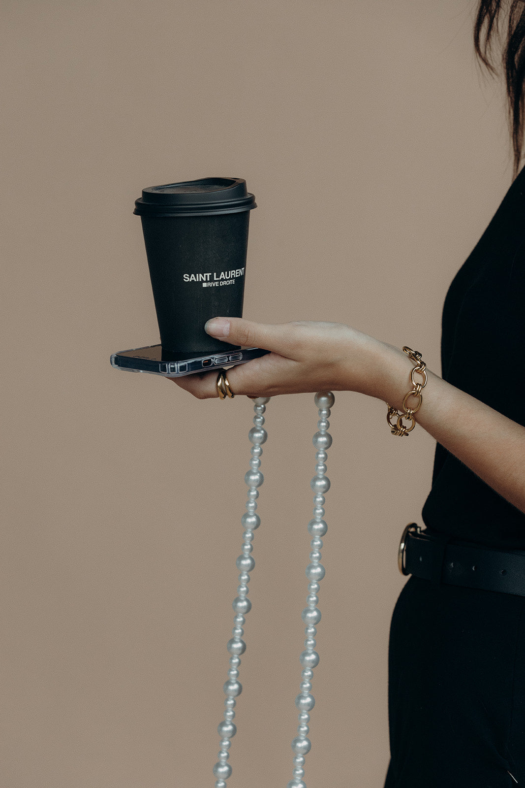 Close-up of a model's hand holding a phone with the Folie à Trois Sabine Pearl wholesale phone chain draped from it and a coffee cup balanced on top.