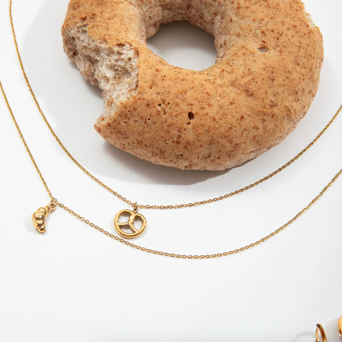 The Salty Sweet NECKLACE and its matching croissant charm necklace draped over a bitten bagel on a white plate.