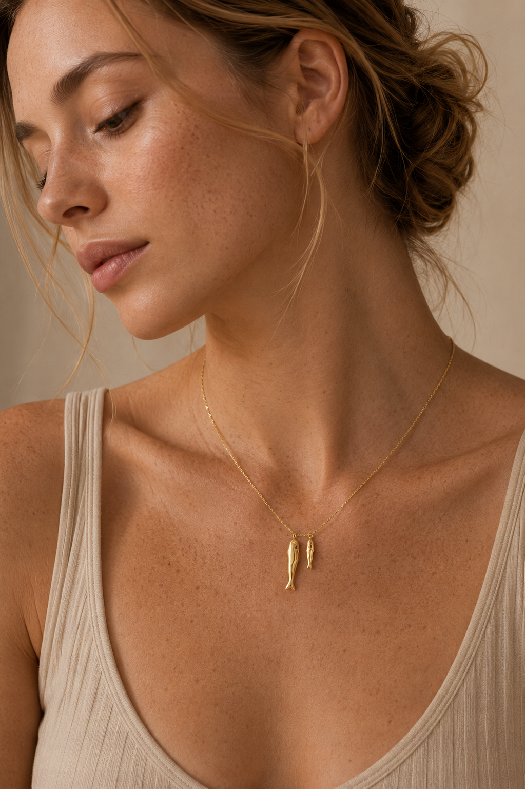 Model wears the gold Sardina NECKLACE with its two fish charms resting at the neckline, over a cream ribbed top.