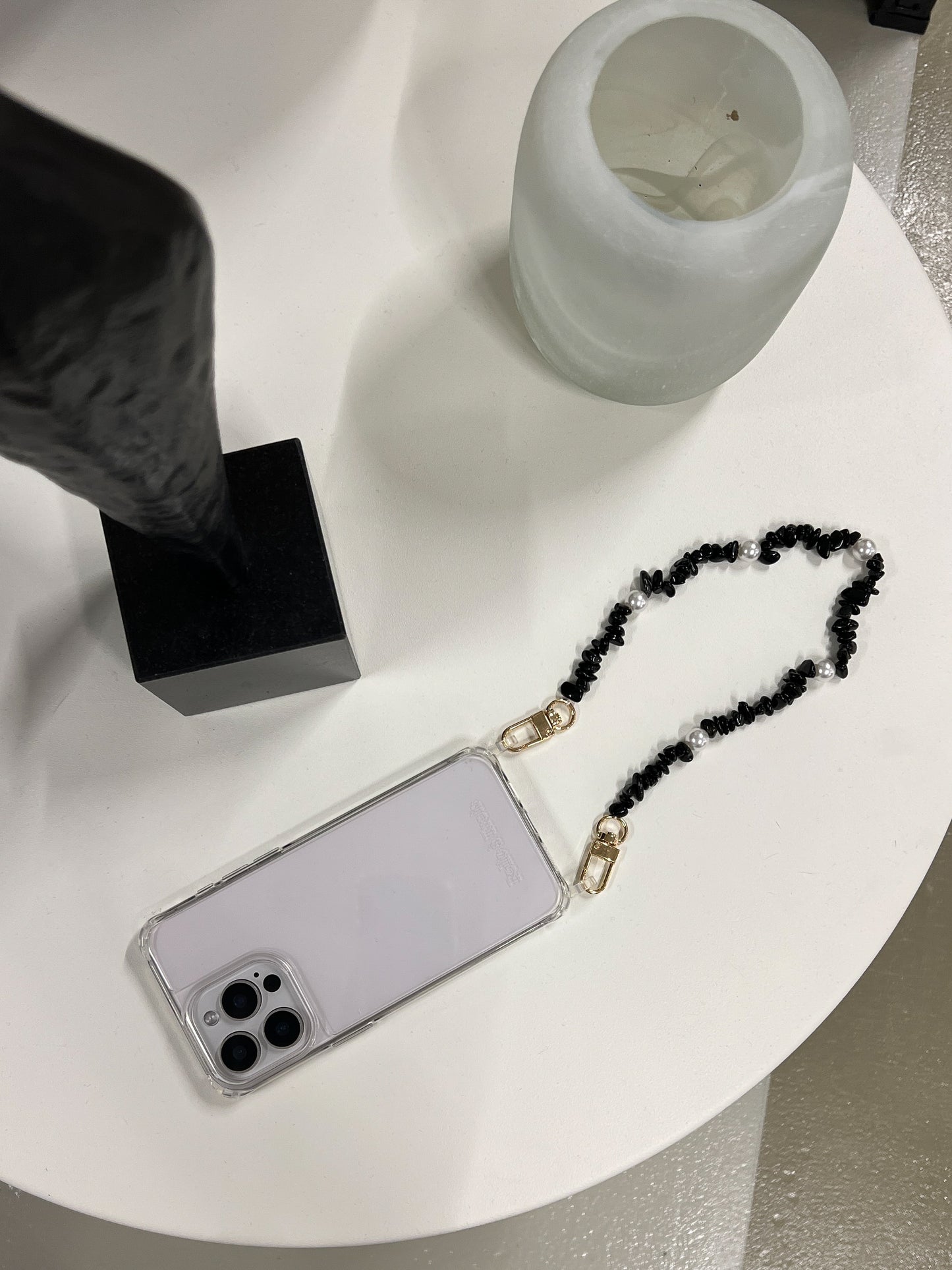 Black beaded Shell Whisper SHORT PHONE STRAP attached to a clear phone case, styled on a white tabletop.
