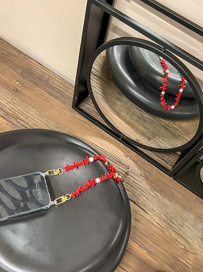 Red beaded Shell Whisper SHORT PHONE STRAP attached to a clear phone case, resting on a black stool with mirror reflection.