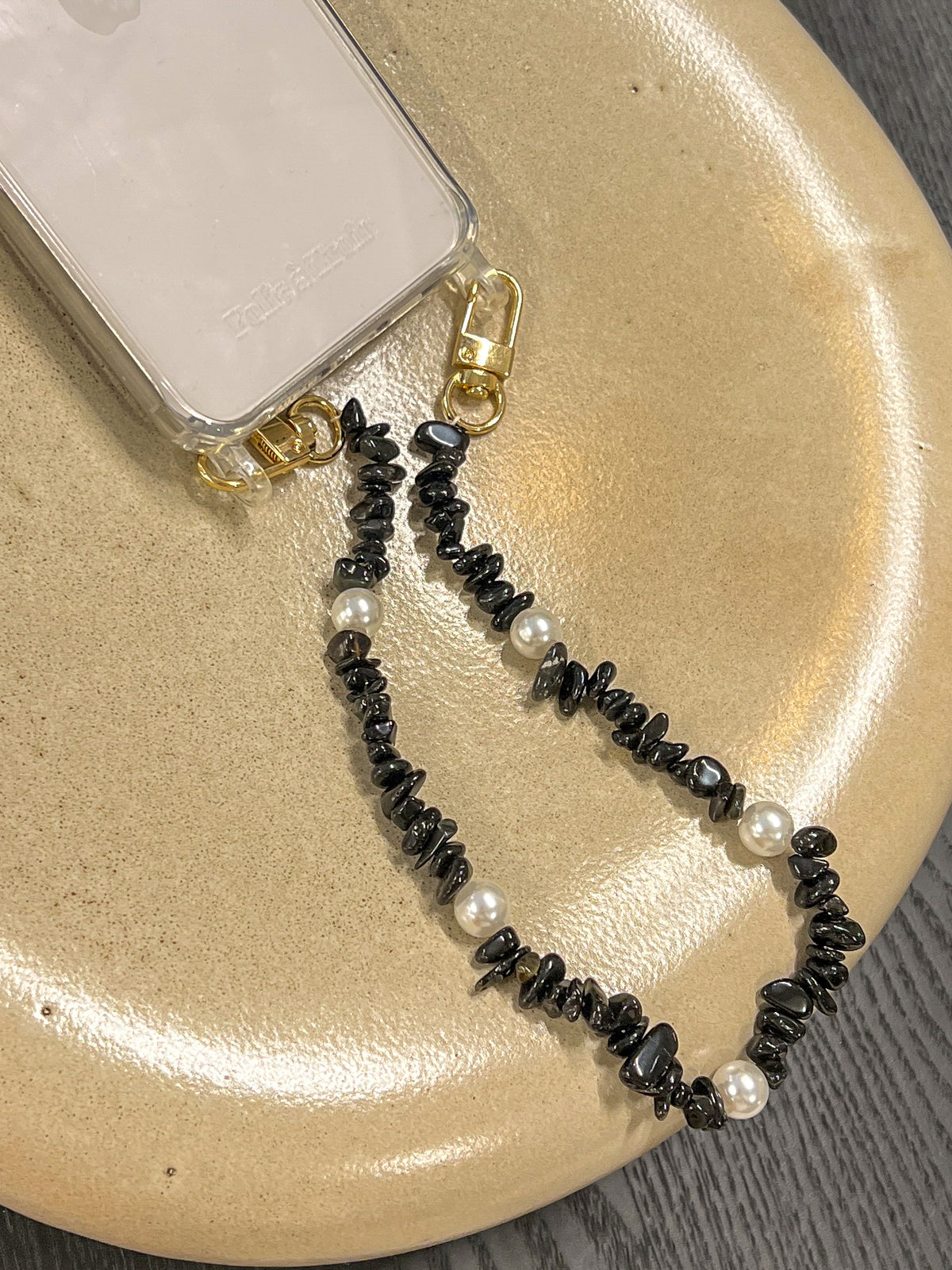 Black beaded Shell Whisper SHORT PHONE STRAP attached to a clear phone case, styled on a tan ceramic plate.