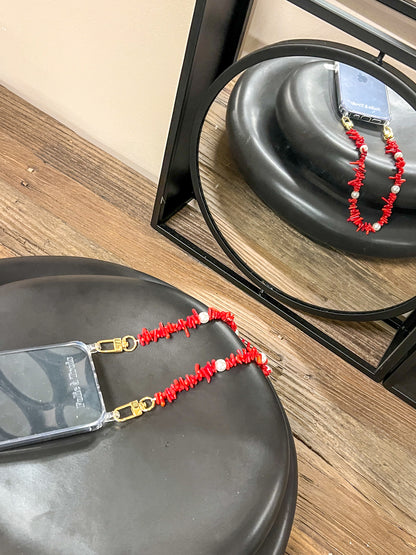 Red beaded Shell Whisper SHORT PHONE STRAP attached to a clear phone case, resting on a black stool near a mirror.