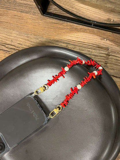 Red beaded Shell Whisper SHORT PHONE STRAP attached to a clear phone case, resting on a black stool on a wood floor.