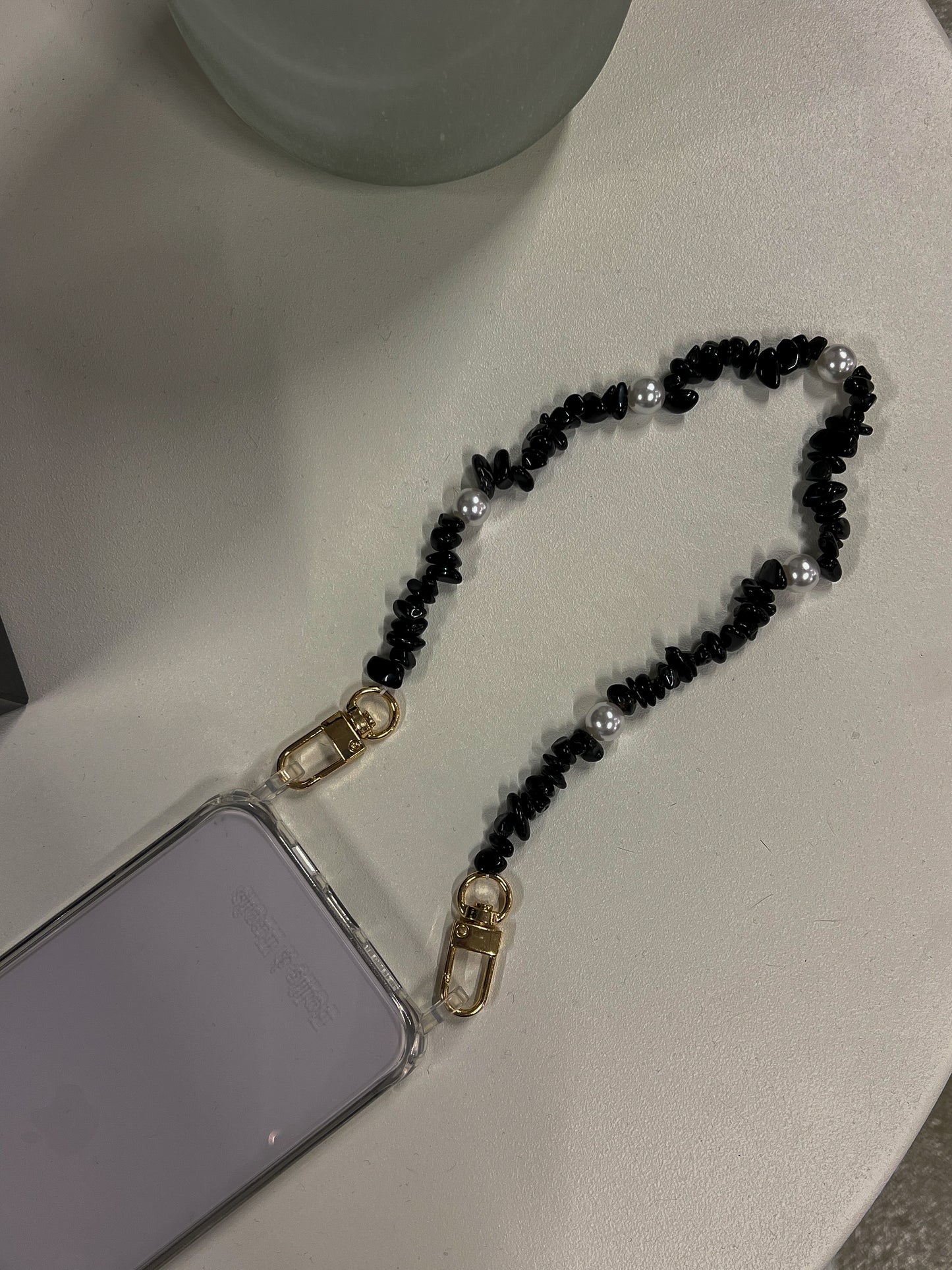 Black beaded Shell Whisper SHORT PHONE STRAP attached to a clear phone case, styled on a white surface.